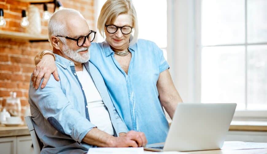 Beautiful senior couple in blue shirts hugging together working with laptop on the kitchen at home