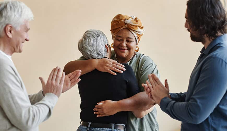 Waist up portrait of diverse group of elderly people applauding and embracing while celebrating success in support group